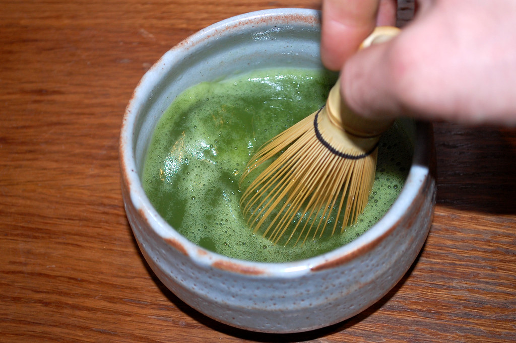 A bamboo whisk stirs vivid green matcha tea in a ceramic bowl