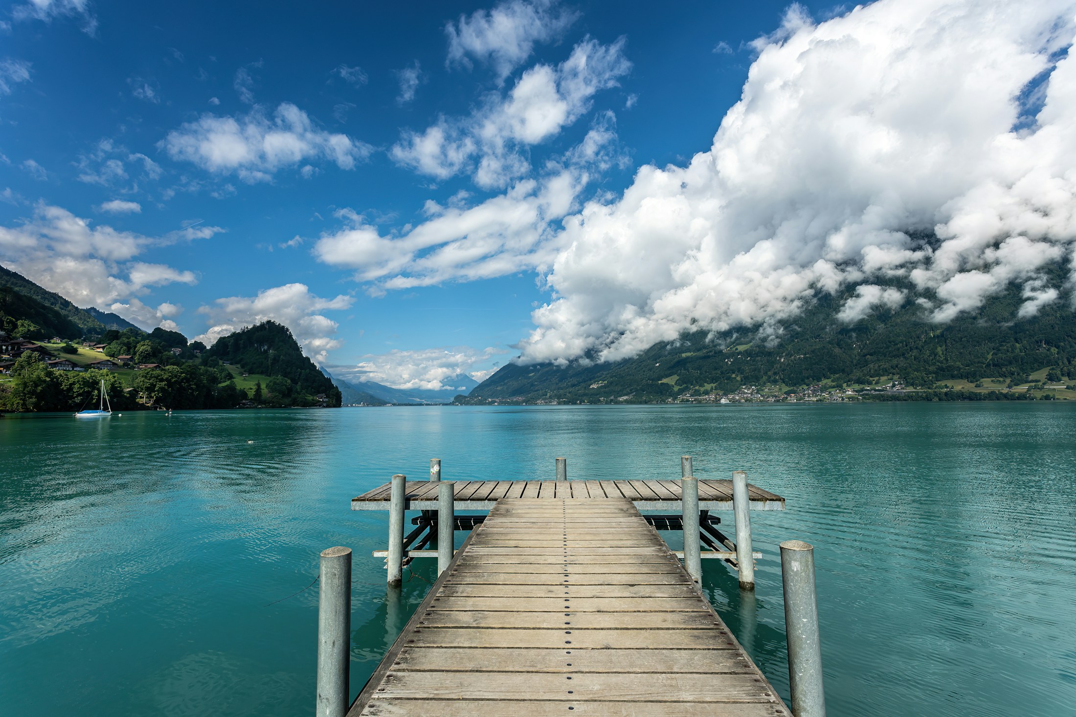 A wooden dock extends into a clear Swiss lake framed by green mountains and clouds