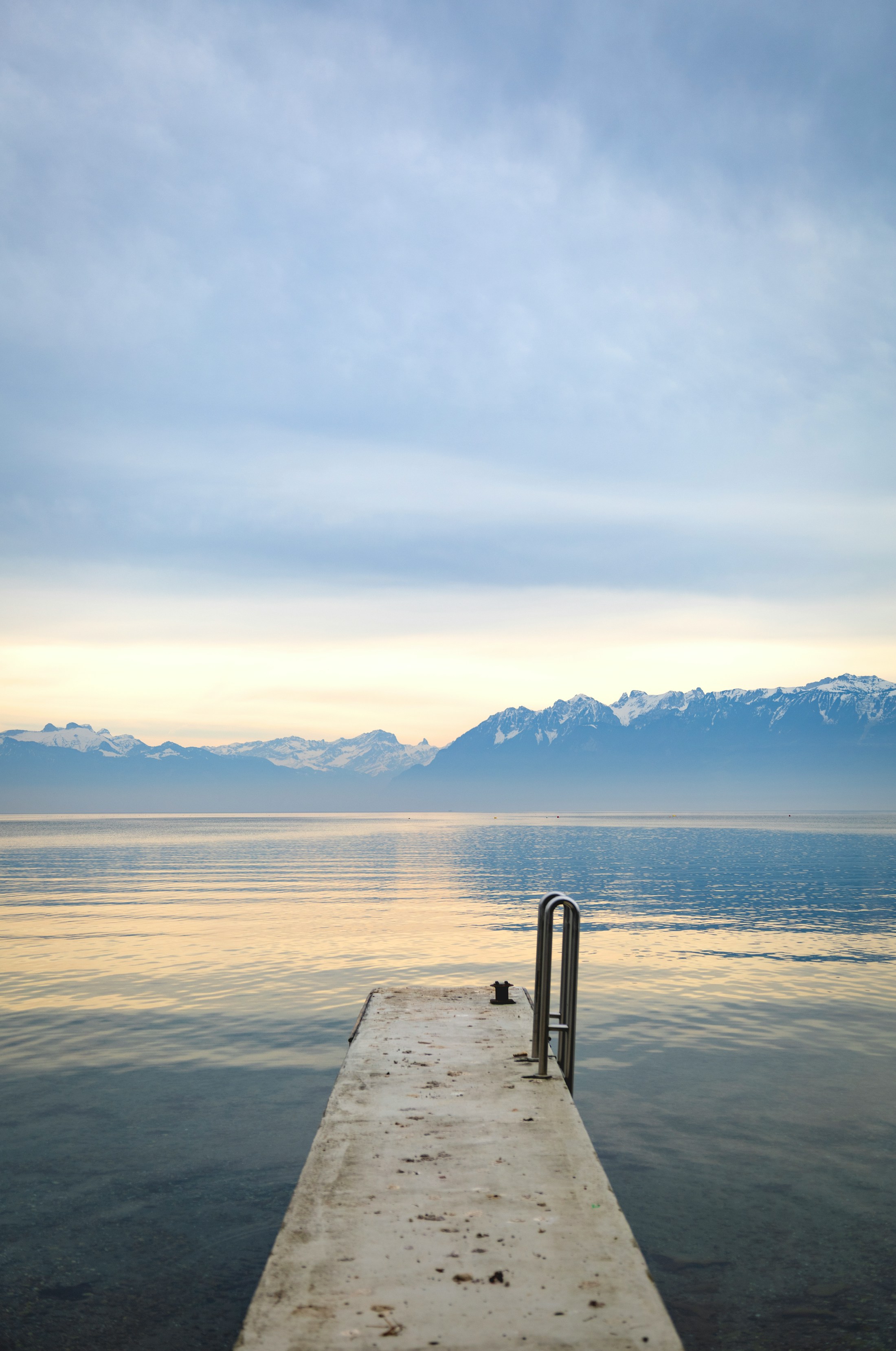 A quiet Swiss lakeside pier points toward snow-dusted mountains at early light