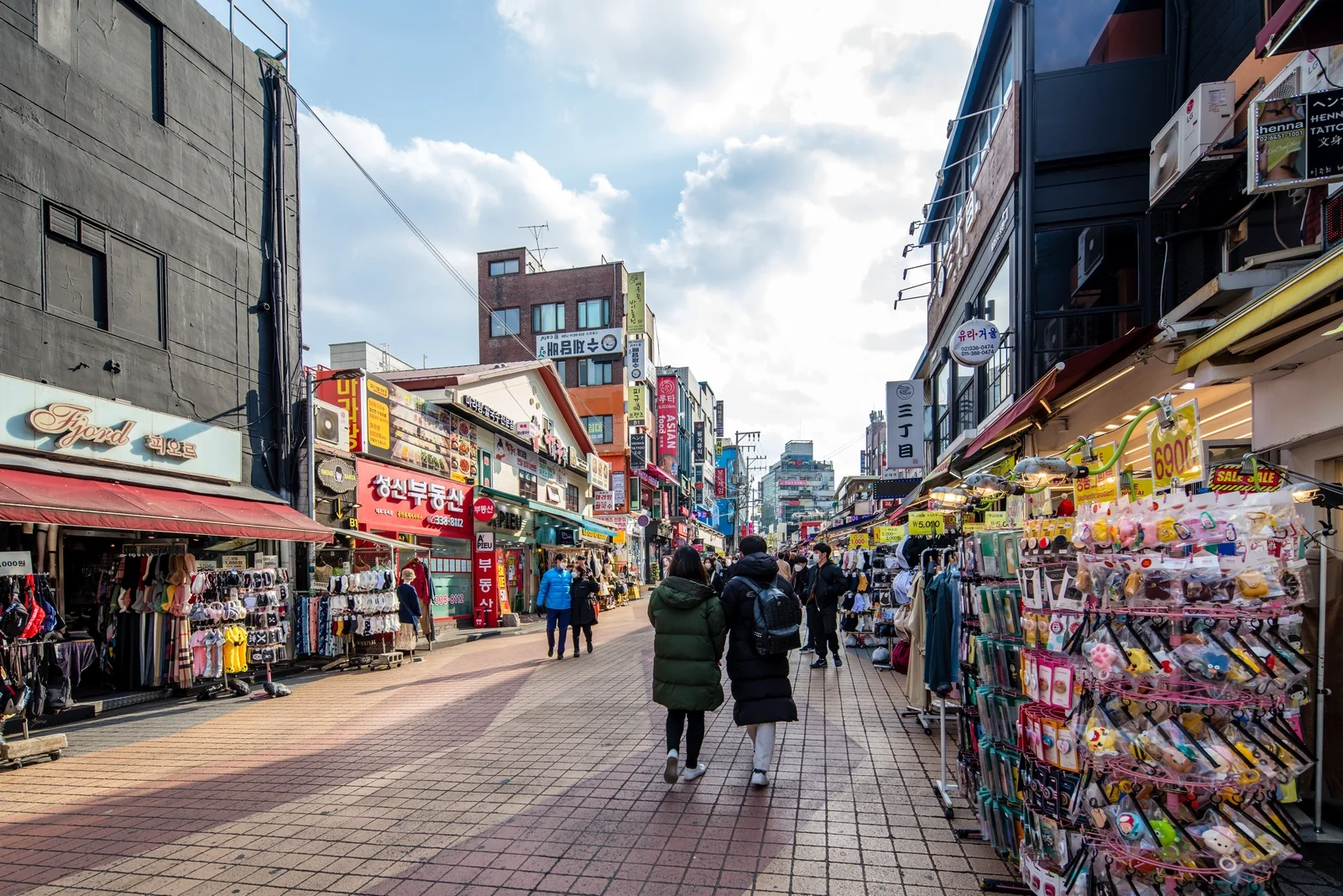 Pedestrians walking between storefronts and outdoor merchandise displays on Hongdae Art Street.