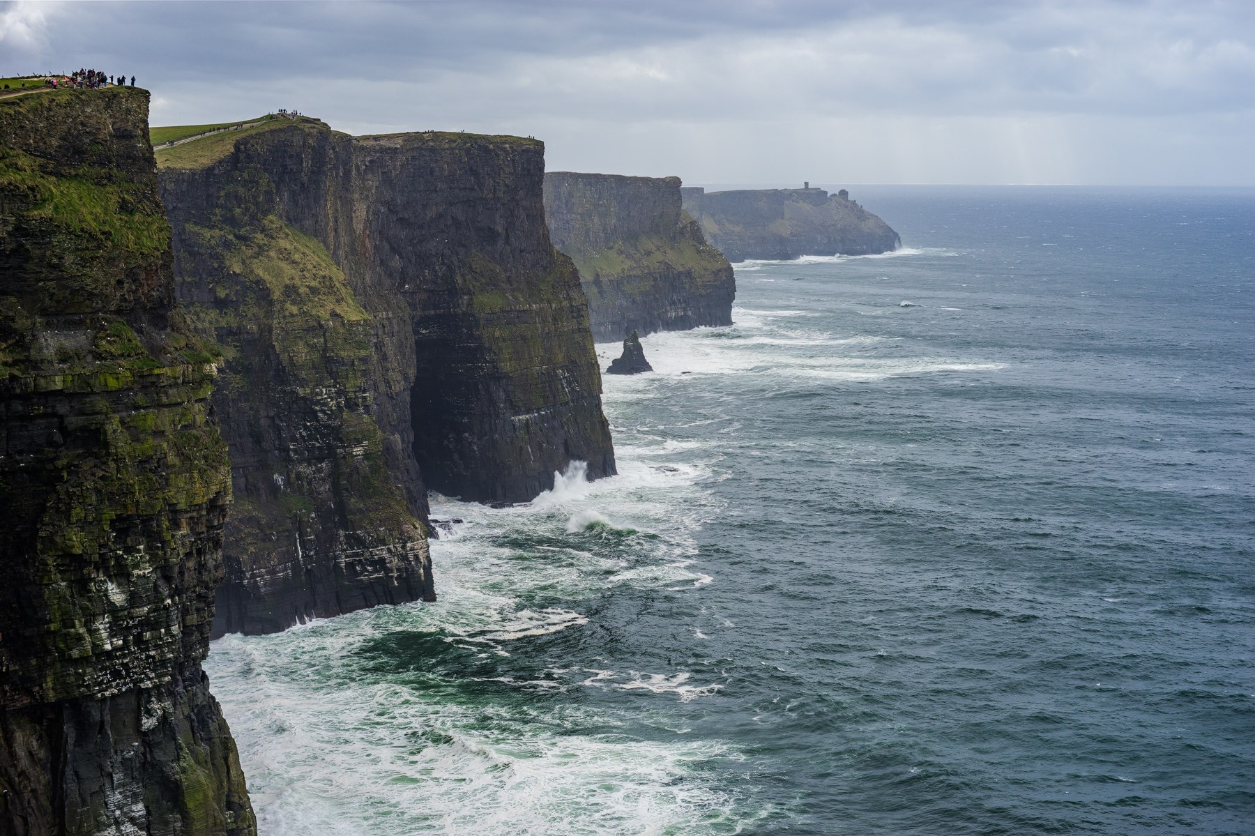 Green-topped Cliffs of Moher descending into Atlantic waves beneath a cloudy Irish sky